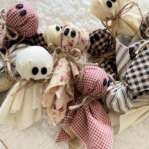 May include: A collection of fabric ghost ornaments featuring various patterns including red and black gingham, floral prints, and solid cream fabric. Each ghost has two black felt circles for eyes and is tied with natural jute twine at the neck.
