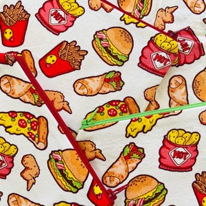 May include: White fabric with a repeating pattern of fast food items, including burgers, fries, pizza, and burritos. Red zippers with a red pull and a green zipper with a green pull are visible. The fries are in red containers with a smiling face.
