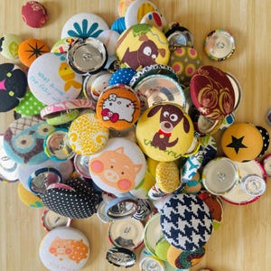 May include: A collection of colorful, handmade buttons with various designs, including cartoon characters, floral patterns, and geometric shapes. The buttons are in different sizes and colors, scattered on a wooden surface.