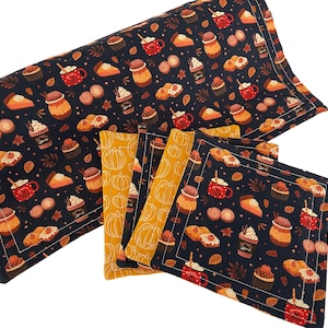 Puede incluir: Un juego de manteles individuales y posavasos azul oscuro con un estampado de temática otoñal. El estampado incluye imágenes de tazas de café, pasteles y hojas en tonos naranja, rojo y marrón. Los posavasos son de color amarillo mostaza con contornos de calabaza blancos.