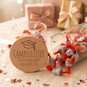 Könnte beinhalten: Ein durchsichtiges Glasgefäß gefüllt mit einzeln verpackten Süßigkeiten, dazu ein runder Holzdeckel mit eingraviertem Doktorhut-Symbol und dem Text CAMPUSFUTTER FÜR HUNGRIGE KÖPFE UND GROBE ZIELE, platziert auf einer hellen, stoffartigen Oberfläche.