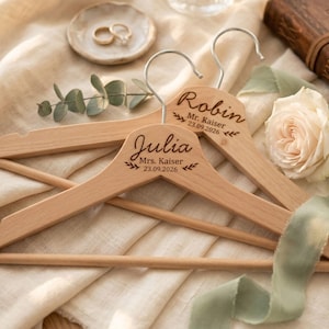 May include: Two wooden clothes hangers engraved with the names "Julia" and "Robin" and a wedding date. The hangers are light brown with silver hooks. A white rose, green ribbon, and rings are also in the image.