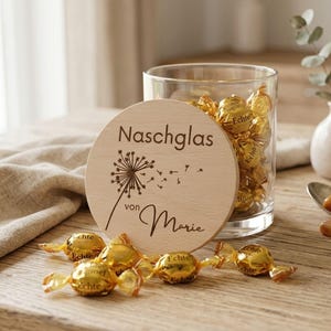 Könnte beinhalten: Ein klares Glas mit goldverpackten Süßigkeiten, neben einem Holzdeckel mit geätztem Text und einem Löwenzahnmotiv. Mehrere Süßigkeiten sind auf der Holzoberfläche verstreut. Eine kleine Vase mit Blumen befindet sich im Hintergrund.