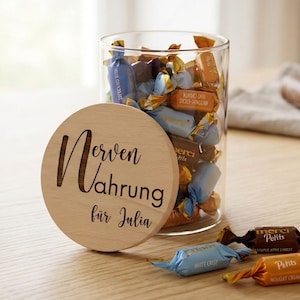Könnte beinhalten: Ein klares Glas mit verpackten Süßigkeiten, mit einem Holzdeckel mit der Aufschrift "Nervennahrung für Julia". Mehrere Süßigkeiten sind auf der Holzoberfläche verstreut. Die Süßigkeiten sind in blauen, orangen und braunen Verpackungen.