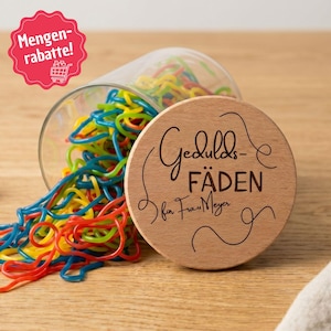 Könnte beinhalten: Ein durchsichtiges Glasgefäß gefüllt mit bunten roten, gelben, blauen und grünen Fruchtgummischnüren. Der runde Holzdeckel ist mit dem Text Gedulds-FÄDEN für Frau Meyer sowie dekorativen geschwungenen Linienmustern graviert.