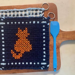 May include: A navy blue and orange cat-themed potholder with a light blue and white potholder and a blue spatula on a wooden cutting board. The cat design is cross-stitched. The potholders have metal hanging rings.