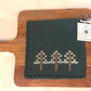 May include: A dark green woven kitchen towel with a cross-stitch tree design, resting on a wooden cutting board. The towel has a tag attached with the text "THE GARDENING WITH CARE SHOP".