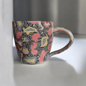 May include: A ceramic mug with a colorful abstract pattern in shades of pink, yellow, and blue on a white background. The mug has a rounded shape and a handle.