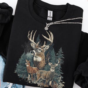 Pu&ograve; includere: Felpa nera con un'illustrazione dettagliata di tre cervi in una foresta. Il design include un grande cervo con corna e due cervi pi&ugrave; piccoli. Una collana d'argento con un pendente circolare &egrave; drappeggiata sulla felpa.