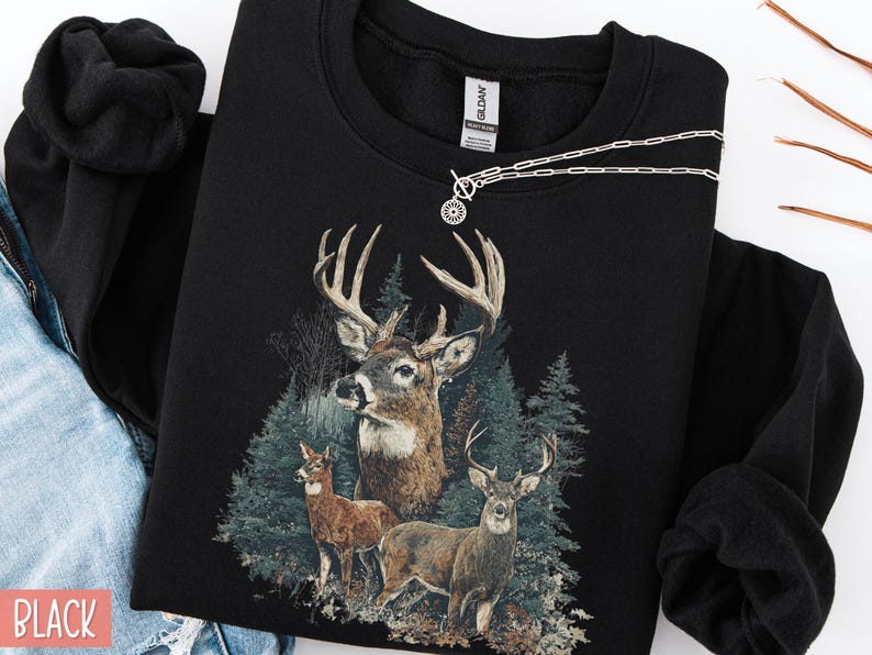 Pu&ograve; includere: Felpa nera con un'illustrazione dettagliata di tre cervi in una foresta. Il design include un grande cervo con corna e due cervi pi&ugrave; piccoli. Una collana d'argento con un pendente circolare &egrave; drappeggiata sulla felpa.