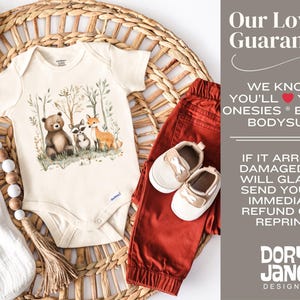 May include: A cream-colored onesie with a watercolor illustration of a bear, raccoon, and fox in a forest setting. The onesie is on a woven wicker surface with a wooden bead garland and a pair of red pants and beige shoes.