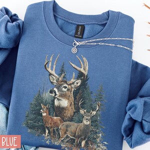 Pu&ograve; includere: Felpa blu indaco con grafica di cervi raffigurante tre cervi in un bosco. La felpa ha girocollo e maniche lunghe. Una collana d'argento con un ciondolo a forma di sole &egrave; drappeggiata sul davanti.