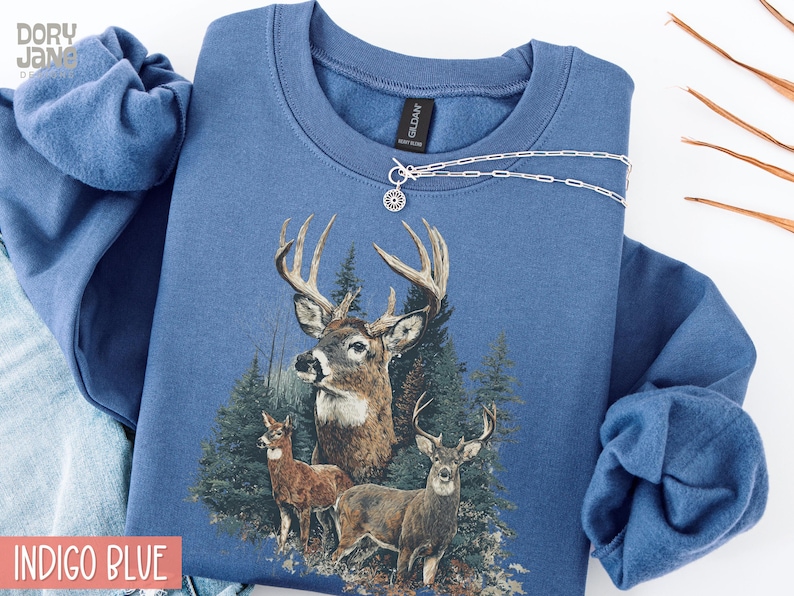 Pu&ograve; includere: Felpa blu indaco con grafica di cervi raffigurante tre cervi in un bosco. La felpa ha girocollo e maniche lunghe. Una collana d'argento con un ciondolo a forma di sole &egrave; drappeggiata sul davanti.