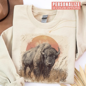 May include: Cream-colored sweatshirt featuring a bison illustration with a sunset backdrop. The design includes the text "Personalize 10 Color Options". The sweatshirt is folded, with a brown hat visible in the background.
