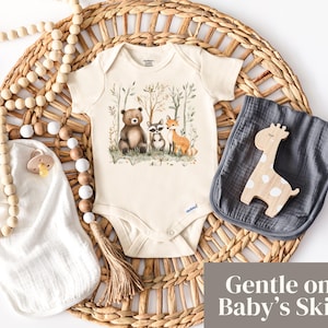 May include: A white baby onesie with a brown bear, raccoon, and fox illustration in a forest setting. The onesie has a brown tassel and the text "Gentle on Baby's Skin".