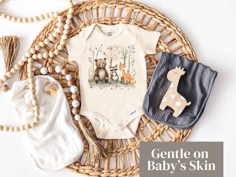 May include: A white baby onesie with a brown bear, raccoon, and fox illustration in a forest setting. The onesie has a brown tassel and the text "Gentle on Baby's Skin".