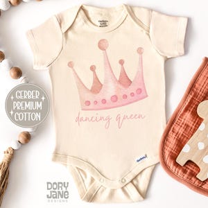 May include: Cream-colored baby bodysuit with a watercolor-style pink crown and the words "dancing queen." The bodysuit is made of Gerber Premium Cotton. A wooden giraffe toy and a rust-colored bib are next to the bodysuit.