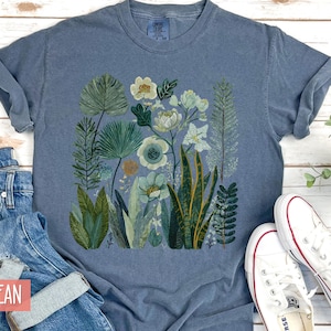 May include: A blue t-shirt with a botanical print of green, white, and blue plants and flowers. The shirt is shown with blue jeans and white trainers.