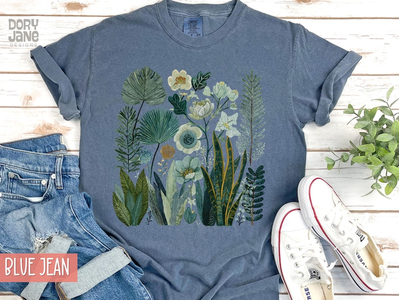 May include: A blue t-shirt with a botanical print of green, white, and blue plants and flowers. The shirt is shown with blue jeans and white trainers.