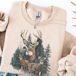 Pu&ograve; includere: Una felpa color sabbia con una grafica di cervi in un ambiente forestale. Il design presenta tre cervi con corna e alberi sempreverdi. Una delicata collana con un ciondolo a forma di sole &egrave; appoggiata sulla felpa.