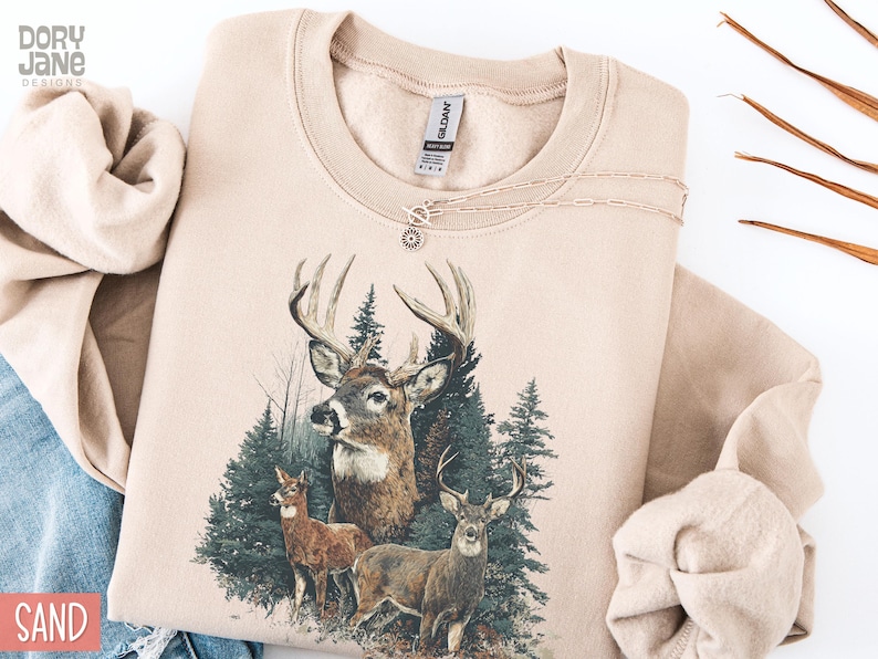 Pu&ograve; includere: Una felpa color sabbia con una grafica di cervi in un ambiente forestale. Il design presenta tre cervi con corna e alberi sempreverdi. Una delicata collana con un ciondolo a forma di sole &egrave; appoggiata sulla felpa.