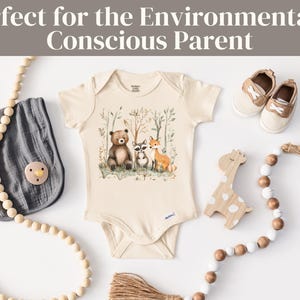 May include: A cream-colored baby onesie with a woodland animal print featuring a bear, raccoon, and fox. The onesie is surrounded by wooden beads and a wooden giraffe toy.