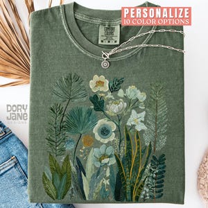 May include: An olive green t-shirt with a floral design featuring white and blue flowers and various green leaves. A silver necklace with a pendant is on the shirt. The text "PERSONALIZE 10 COLOR OPTIONS" is at the top.