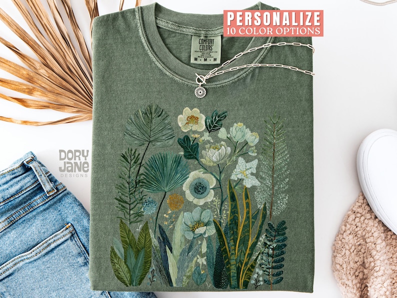 May include: An olive green t-shirt with a floral design featuring white and blue flowers and various green leaves. A silver necklace with a pendant is on the shirt. The text "PERSONALIZE 10 COLOR OPTIONS" is at the top.