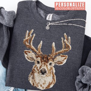 May include: Dark gray sweatshirt featuring an embroidered deer head with brown and beige details. The sweatshirt has a crew neck and long sleeves. A silver necklace with a circular pendant is draped across the front. Text at the top reads "PERSONALIZE 10 COLOR OPTIONS."