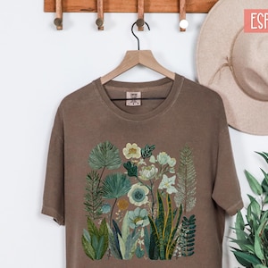 May include: Espresso-coloured t-shirt with a floral design featuring various green and blue plants and flowers. The shirt is hanging on a wooden hanger. The text "Dory Jane Designs" is visible on the bottom left.