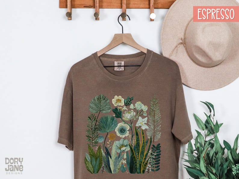 May include: Espresso-coloured t-shirt with a floral design featuring various green and blue plants and flowers. The shirt is hanging on a wooden hanger. The text "Dory Jane Designs" is visible on the bottom left.