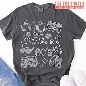 Puede incluir: Una camiseta gris oscuro con un dise&ntilde;o gr&aacute;fico blanco que presenta iconos de los 80 como una radiocassette, patines, una cinta de casete y gafas de sol. El texto "I love the 80's 80's" est&aacute; impreso en la camiseta.