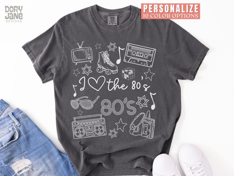 Puede incluir: Una camiseta gris oscuro con un dise&ntilde;o gr&aacute;fico blanco que presenta iconos de los 80 como una radiocassette, patines, una cinta de casete y gafas de sol. El texto "I love the 80's 80's" est&aacute; impreso en la camiseta.
