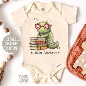 May include: Cream-colored baby onesie with a cartoon illustration of a green caterpillar wearing glasses and leaning on a stack of books. The text "future bookworm" is below the illustration. A wooden giraffe and a bib are on the right.