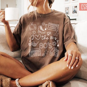 Puede incluir: Una camiseta marr&oacute;n con un dise&ntilde;o gr&aacute;fico blanco que presenta una cinta de casete, patines, una radiograbadora, un televisor y el texto "I love the 80's 80's".