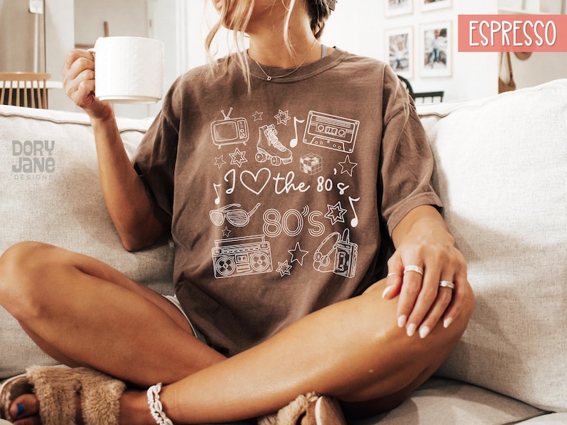 Puede incluir: Una camiseta marr&oacute;n con un dise&ntilde;o gr&aacute;fico blanco que presenta una cinta de casete, patines, una radiograbadora, un televisor y el texto "I love the 80's 80's".