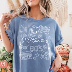 Puede incluir: Una camiseta azul vaquero con un dise&ntilde;o gr&aacute;fico blanco que incluye una cinta de casete, patines, un televisor, gafas de sol, una radiograbadora, auriculares y estrellas. El texto de la camiseta dice "I love the 80's 80's".