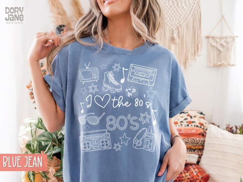 Puede incluir: Una camiseta azul vaquero con un dise&ntilde;o gr&aacute;fico blanco que incluye una cinta de casete, patines, un televisor, gafas de sol, una radiograbadora, auriculares y estrellas. El texto de la camiseta dice "I love the 80's 80's".