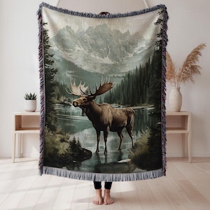 May include: A woven throw blanket featuring a scenic landscape with a moose standing in a lake. The design includes a mountain range, evergreen trees, and a body of water. The blanket has a fringed edge and is primarily in shades of green, brown, and gray.