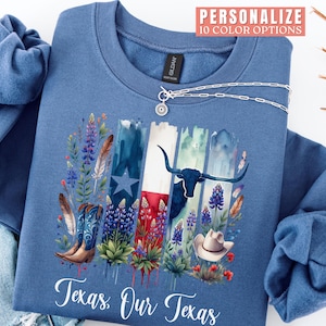 May include: A blue sweatshirt featuring a Texas-themed design. The graphic includes the Texas flag, a longhorn, cowboy boots, a cowboy hat, and wildflowers. The text "Texas Our Texas" is at the bottom. A silver necklace is draped across the sweatshirt.