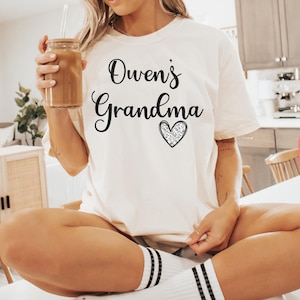 May include: A white t-shirt with the text "Owen's Grandma" and a sparkly heart. The shirt is on a person sitting on a chair.