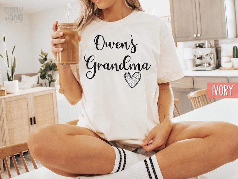 May include: A white t-shirt with the text "Owen's Grandma" and a sparkly heart. The shirt is on a person sitting on a chair.