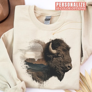 May include: Cream-colored sweatshirt featuring a detailed illustration of a bison's head, with a landscape overlay. The design incorporates shades of brown and gray. The text "PERSONALIZE 10 COLOR OPTIONS" is visible.