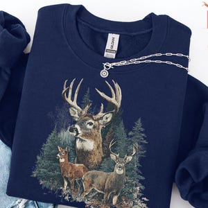 Pu&ograve; includere: Felpa blu navy con grafica dettagliata di tre cervi in un ambiente forestale. Il design include un grande cervo con corna imponenti e due cervi pi&ugrave; piccoli. Una collana d'argento con un ciondolo circolare &egrave; drappeggiata sulla felpa.