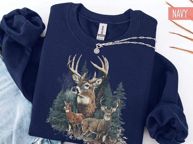 Pu&ograve; includere: Felpa blu navy con grafica dettagliata di tre cervi in un ambiente forestale. Il design include un grande cervo con corna imponenti e due cervi pi&ugrave; piccoli. Una collana d'argento con un ciondolo circolare &egrave; drappeggiata sulla felpa.