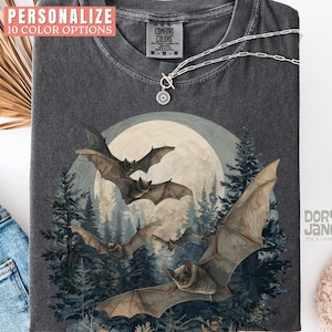 May include: Dark grey t-shirt with a watercolour illustration of bats flying in front of a full moon and a forest. The shirt has the text "PERSONALIZE 10 COLOR OPTIONS" at the top. A silver necklace is draped across the shirt.