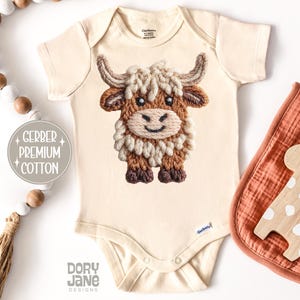 Pode incluir: Body de beb&eacute; creme com um design bordado detalhado de uma vaca das Terras Altas. A vaca apresenta pelo castanho e creme, um rosto sorridente e pequenos chifres. O body tem o texto "Gerber Premium Cotton" e "Dory Jane Designs".