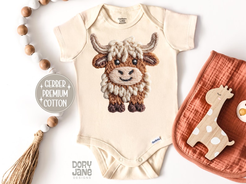 Pode incluir: Body de beb&eacute; creme com um design bordado detalhado de uma vaca das Terras Altas. A vaca apresenta pelo castanho e creme, um rosto sorridente e pequenos chifres. O body tem o texto "Gerber Premium Cotton" e "Dory Jane Designs".