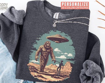May include: Dark gray sweatshirt featuring a graphic of Bigfoot, an alien, and a UFO over a desert landscape. The design includes the text "PERSONALIZE 10 COLOR OPTIONS" in a red banner. A silver necklace is draped over the sweatshirt.
