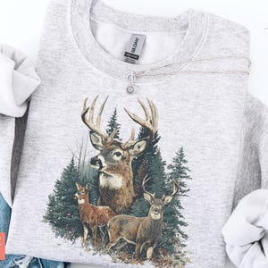 Pu&ograve; includere: Felpa color cenere con una grafica dettagliata di tre cervi in un ambiente forestale. Il design include un grande cervo con corna, due cervi pi&ugrave; piccoli e alberi sempreverdi. Le parole "SUPER COZY" sono stampate in basso a sinistra.